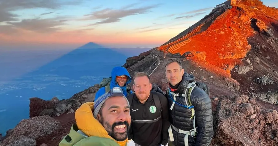 Drei lächelnde Wanderer am roten Vulkanhang des Fuji bei Sonnenaufgang, der Schatten des Berges fällt auf die Wolken